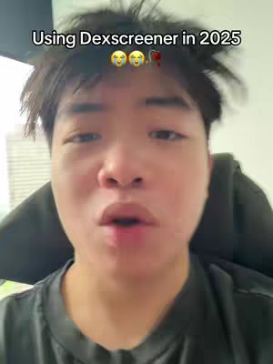 A selfie-style video cover image featuring a young man with a soft, slightly out-of-focus background. The man has short, dark hair and is looking directly at the camera with a slight expression of concern or surprise. Above his head, a text overlay reads, 'Using Dexscreener in 2025' followed by relevant emoji. The lighting appears to be natural, possibly from a window, as the image lacks harsh shadows. The overall composition is simple, focusing intently on the subject's face with some blurred details surrounding him.