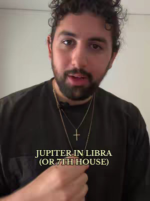 A man with dark curly hair and a beard is centered in a close-up shot, looking towards the camera. He's wearing a black t-shirt with a thin gold necklace and a cross pendant.