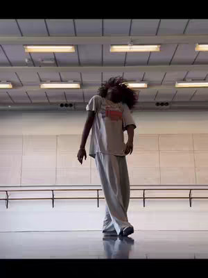 The image captures a person in a dance studio, possibly in motion. The individual, likely female, is seen mid-dance, with her hair in movement. She is wearing a casual outfit, and the studio setting with long lighting fixtures overhead is visible. The composition is dynamic, with the focus on movement.