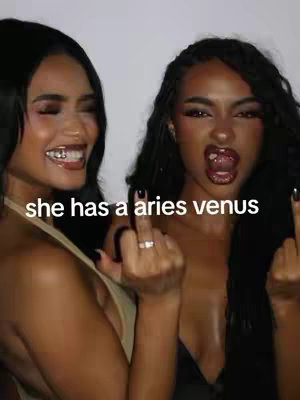 Two women pose side-by-side, one on the left smiling with silver teeth and the other on the right with her tongue out, both holding up the middle finger. The text overlay reads 'she has a aries venus'.