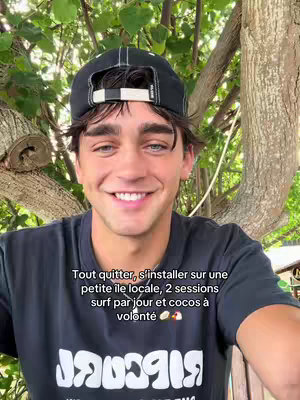 A young man with brown hair and a black cap smiles at the camera. He is in front of a backdrop of green leaves and branches.