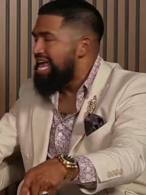 A man with a well-groomed beard and haircut is seen in a close-up shot, wearing a patterned shirt under a cream-colored blazer. He is raising his head and opening his mouth as if speaking passionately, with a gold watch and ring adding detail to his appearance.