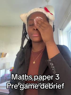 A young Black woman is shown with her eyes closed pressing a makeup pad to her left eye, she is also wearing a white cat ear headband with a small red bow, a gold necklace, and a dark blazer over a brown shirt. Text at the bottom reads 'Maths paper 3 Pre game debrief'.