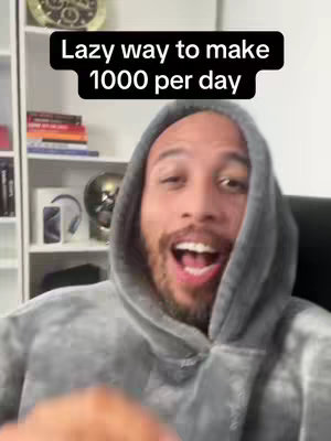 A selfie-style video cover featuring a man in a gray hoodie. The man is smiling widely with an open mouth. A text overlay in a black box reads \"Lazy way to make 1000 per day\". The background includes a bookshelf with books, decor, and an out-of-focus glimpse of a smartphone.