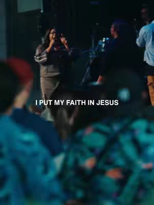 A person is singing on stage at a concert, microphone in hand. Text overlayed on the middle of the image says, 'I put my faith in Jesus'. There are blurred figures in the background.
