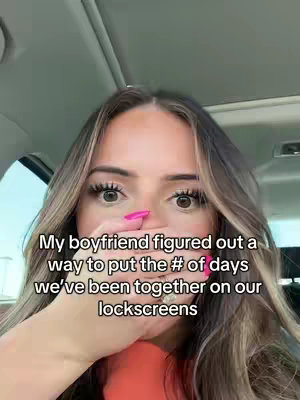 A woman with long blonde hair is shown inside of a car. She is holding her hand over her mouth and wearing pink nail polish. Text overlays read: "My boyfriend figured out a way to put the # of days we've been together on our lockscreens."