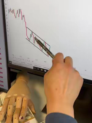 The image shows a person's hands pointing at a stock chart on a computer screen. A wooden hand is seen holding money. The color palette is mainly white, with red and green lines on the chart, and the overall composition is about trading.