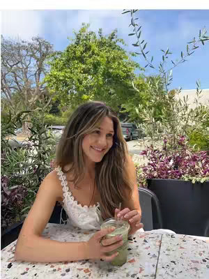A woman is seated at a table outdoors, smiling joyfully while holding a tall, green drink. She is wearing a white top, with her long hair framing her face. The setting includes a table with a patterned top and greenery in the background.