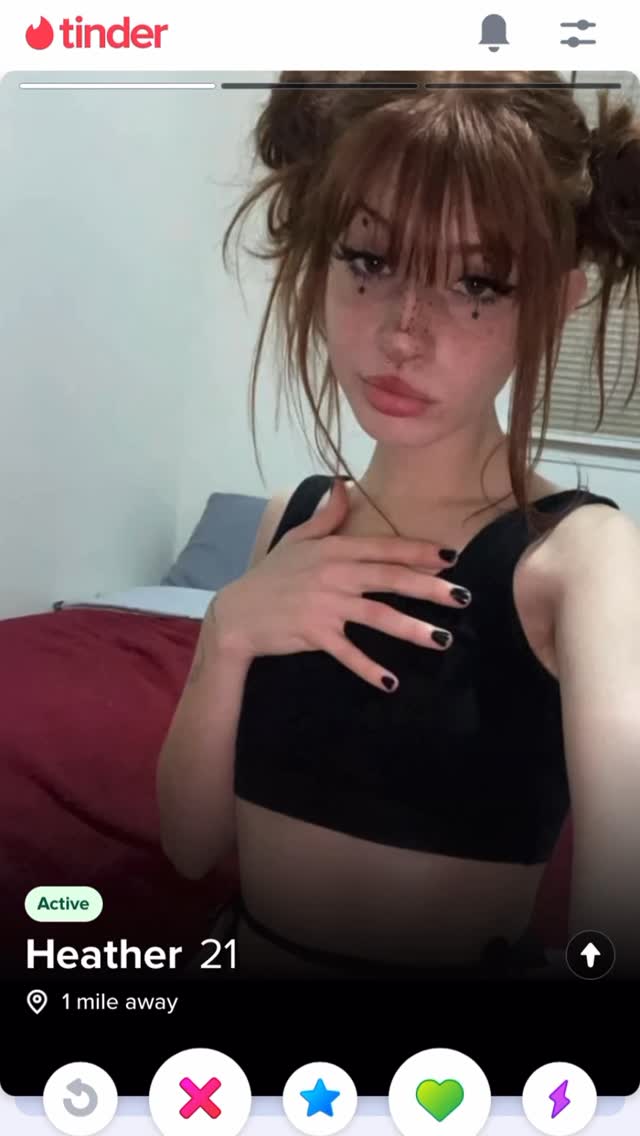 A woman with reddish-brown hair and two buns, makeup with dark eyeliner and freckles drawn on, is on a social media app presumably Tinder. The background includes a white wall, a bed with a red comforter, a grey pillow. The main color palette of the image is white, red, and the woman has dark black manicured nails on her hand. The app has a white progress bar and the woman is active, 1 mile away.