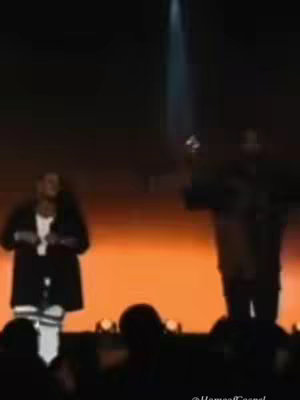 The cover image shows a live performance on a stage, with the background gradient lighting of gold and orange tones. The stage is partially visible at the bottom. A person is on the left as if they are holding a microphone, they are wearing a blazer over a shirt and white pants. The second person on the right is in a dark coat and the artist or performer appears to be raising their arm. A distinct spotlight shines down. Silhouettes of people are in the foreground, likely the audience.
