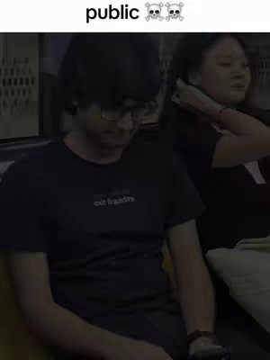 A dimly lit interior shot featuring two individuals. The foreground is dominated by a man wearing glasses and a black t-shirt with white text. To his right, a woman is visible, partially obscured, with her hand near her hair. The word "public" and skull emojis are in the upper center.