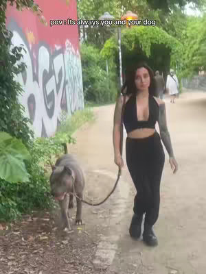 A woman in a black halter top and pants walks with a dog on a leash along a path. The path is bordered by green foliage and a wall with graffiti. In the background, other people are walking.