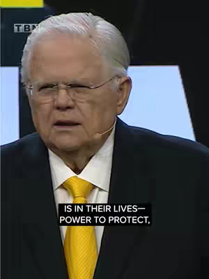 A medium shot features an older man with gray hair, glasses, and a black suit with a yellow tie. The man is speaking, and the background has a blurred appearance with a TBN logo visible.