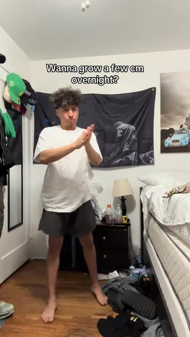 A young man stands center frame in a messy bedroom, clapping his hands together. He is wearing a white t-shirt and black shorts. The background includes a bed, a nightstand, a flag with a black and white image, a mirror, and various items scattered on the floor. Text overlay reads, "Wanna grow a few cm overnight?"