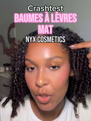 A selfie-style image focusing on a woman's face. She has dark, curly hair and is wearing a white top. The background is a plain wall. Overlay text in pink and white across the top suggests a crash test of matte lip balms from NYX Cosmetics.