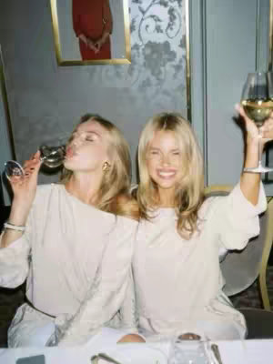 Two young women are seated at a table, likely indoors, with a beige tablecloth and a decorated floral-print wall visible. Both are blonde and wearing light-colored, matching outfits. One woman is tilting her head back to drink from a wine glass while the other is smiling, holding her glass aloft. The lighting indicates a casual, celebratory atmosphere.
