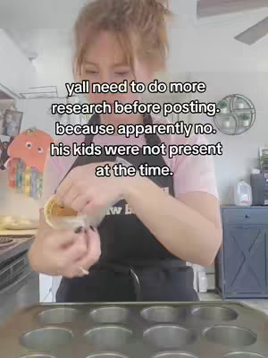 A woman in a pink shirt and black apron is preparing food in a kitchen. She is holding a small container and pouring its contents into a muffin tin. The kitchen has light-colored cabinets and walls, with various kitchen items visible in the background.