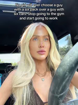 A selfie-style shot from within a car. A woman with long, blonde hair and light skin is in the driver's seat, looking slightly to the side and appearing contemplative. She's wearing a brown top, and multiple necklaces, the steering wheel is visible, and the upper part of the car's interior is dark. The background outside the car is blurred, suggesting motion.