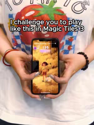 A person holding a smartphone in their hands, a mobile game is being played on it. Overlayed on the image is text asking, "I challenge you to play like this in Magic Tiles 3". In the background over the person's chest and chest is a white t-shirt with a strawberry graphic.