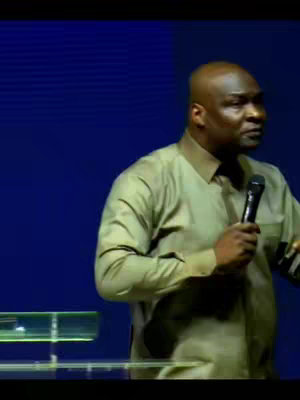 The image shows a medium shot of a bald Black man indoors. He is wearing a light green shirt. He is holding a microphone in his right hand and is gesturing with his left hand, as if he's speaking. A blue background appears behind him.