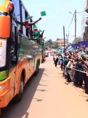 The image shows a sunny street scene with a bus and people gathered. The bus is a large vehicle with an orange and green design, and people are waving from the windows. On the sidewalk, many people are gathered, waving flags and cheering. The atmosphere appears to be celebratory.