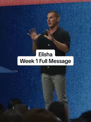 A middle-aged man in a black shirt and gray pants stands on a stage in front of a blue backdrop. The man is gesturing with his hands as the text overlay on the screen reads "Elisha Week 1 Full Message."