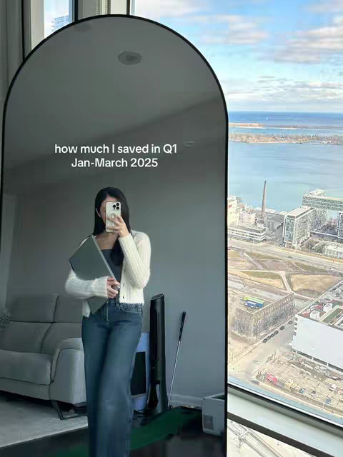 A woman stands in a well-lit room, holding a laptop and a smartphone. The image is a reflection in a large mirror that fills a significant portion of the frame, with the woman positioned centrally. The room has a cityscape view visible and a gray couch.
