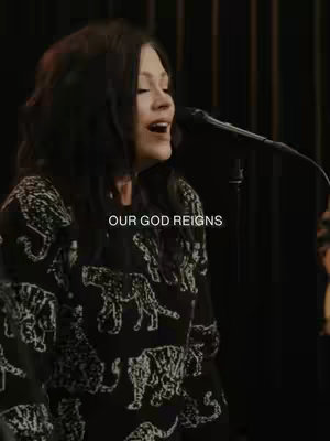 A woman with long dark hair sings into a microphone on a dimly lit stage. She wears a black sweater with a pattern of white tigers. The text 'OUR GOD REIGNS' is overlaid.