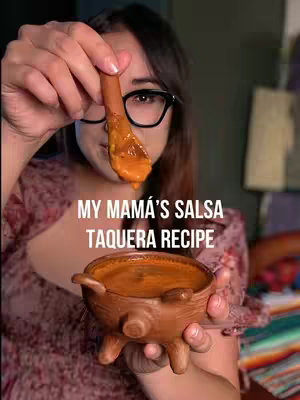 A woman wearing glasses holds a wooden spoon with salsa above a small, decorative bowl filled with salsa. The woman and the bowl are in the foreground, with a dark background. Text overlayed on the image reads: 'MY MAMÁ'S SALSA TAQUERA RECIPE'.