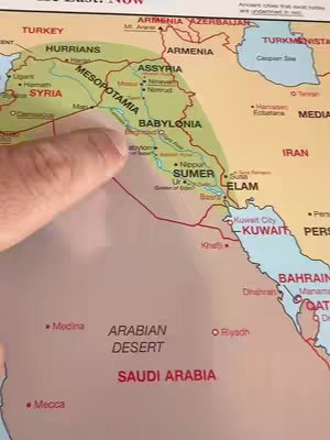 A person's finger points toward a map showing the Middle East. The map has text labels like 'MESOPOTAMIA', 'BABYLONIA', and 'SUMER'. The background appears to be a wooden surface.