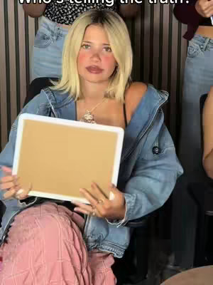 A young woman with blonde hair and a denim jacket is seated, holding a blank white board. She is facing the camera, appearing to be the focal point of the image. Other people are visible in the background. The overall color scheme is warm, with natural skin tones and the neutral white of the board.
