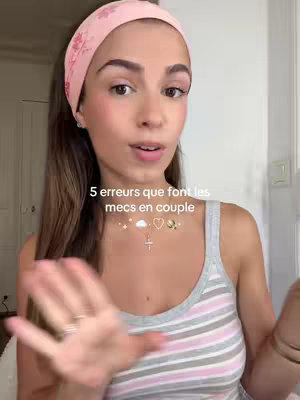 A young woman with long brown hair and a pink floral headband is centered in the frame, looking directly at the camera. The background appears to be a room with a neutral-colored wall and a partially visible door. She is wearing a striped tank top and her hands are gesturing towards the right. Above her, overlaid text reads '5 erreurs que font les mecs en couple', accompanied by decorative icons.