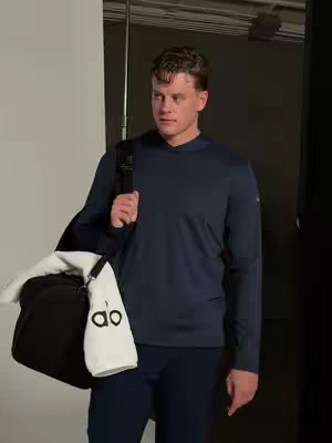 A medium shot of a man standing in a studio setting, possibly a photoshoot. The man is wearing dark blue athletic wear and carrying a black gym bag over his shoulder. A towel with the "alo" logo hangs from the bag. The background is blurred, with a light source visible to the left.