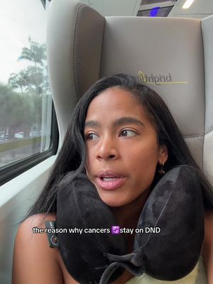 A woman with long dark hair sits in a train seat near a window. She wears a black neck pillow and appears to be looking out the window. The text overlay reads, 'the reason why cancers ♋ stay on DND'.