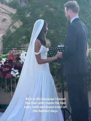 A wedding scene with a bride in a white gown and veil, holding hands with the groom. Lush greenery and floral arrangements provide the backdrop.