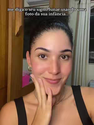 A woman is centrally framed, gazing towards the camera with a slight smile, her hand delicately touching her face near her cheek. She has dark hair pulled back, dark eyes, and neutral makeup, creating a close-up focus. The background subtly shows a wooden cabinet and blurred elements suggesting an indoor setting.