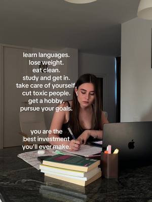 A young woman is sitting at a kitchen counter, writing in a notebook with a pen. There is a laptop, books, and a pen holder on the counter. Text overlays are written in white on the left, including the phrases: 'learn languages. lose weight. eat clean. study and get in. take care of yourself, cut toxic people. get a hobby. pursue your goals. you are the best investment you'll ever make.'