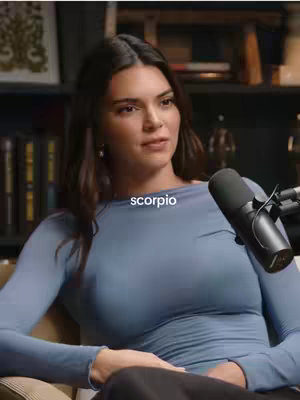 A woman with dark hair and a light blue top sits in front of a muted background. A microphone is positioned to the right of her face. Text "scorpio" is overlaid on the image.