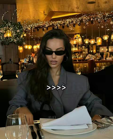 A woman wearing sunglasses and a blazer is seated at a restaurant table, with a restaurant interior visible in the background. The hook text '>>>>' is overlaid on the image.