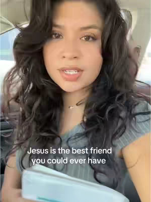 A woman with dark curly hair is in a car. She has a fair complexion and is looking toward the camera speaking. Text overlay says, 'Jesus is the best friend you could ever have'. She is wearing a light-colored top and a necklace. She is holding a white object in her hands.