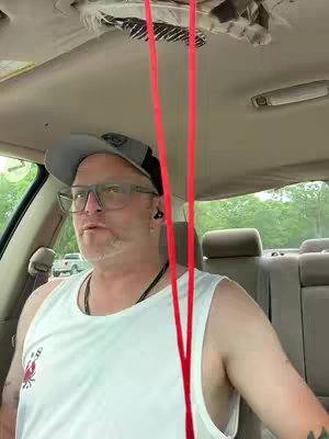 A man wearing glasses and a hat is sitting in the driver's seat of a car, looking slightly to the side. Red straps drape down, possibly from the sun visor area. The interior of the car is visible, with the dashboard and seats in view, and trees out the window.