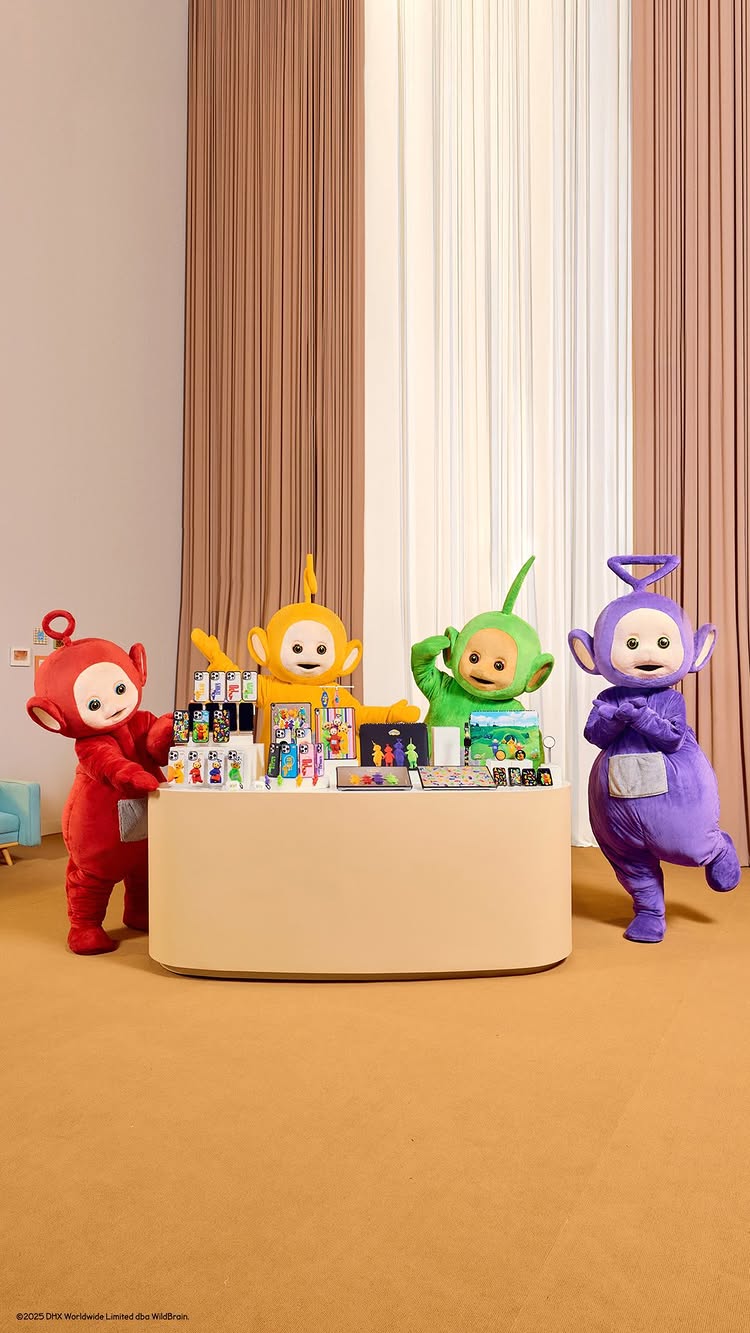 The cover image features the Teletubbies standing behind a display table filled with merchandise. There's a central background with beige, brown, and white vertical stripes, and a plain light-colored wall. The Teletubbies are in their characteristic colors (red, yellow, green, and purple). Merchandise includes phone cases, and other items.