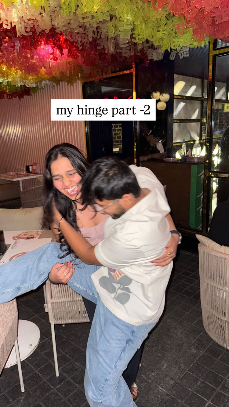 A candid photo captures a moment of joy between a woman and a man in an indoor setting with a restaurant vibe. The woman with long dark hair is laughing heartily while the man is holding the woman in a romantic, fun-filled hug. She is wearing a pink top and light blue jeans, and he's dressed in a white tee and blue jeans. They are in front of a background featuring dangling decorations.