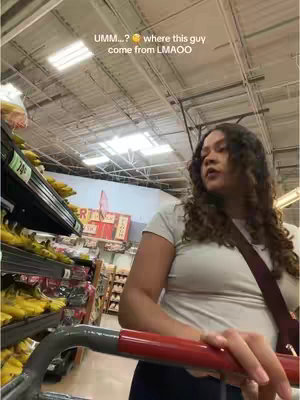 A woman with curly dark hair stands in a grocery store, looking slightly off-center to the right. She's holding a red shopping cart. The background shows shelves of bananas, other produce, and the store's ceiling. Text overlays the image, describing the scene's context.