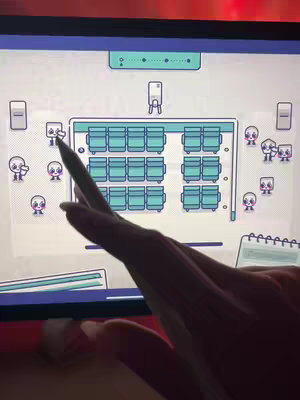 The cover image shows a mobile game interface on a tablet. The game presents a top-down view of an imagined movie theater or auditorium, with rows of teal seats. Stylized cartoon characters are shown in the game, and a finger with a stylus is shown interacting with the screen. The color balance is teal, white and red.