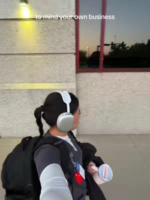 A woman with dark hair in a ponytail is walking away from the camera. She wears a gray scrub top, a black backpack, and white headphones. The background includes a building with a light fixture and a glass window reflecting the sunset.