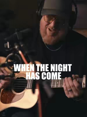 The image shows a man with a beard, glasses, and a hat, holding a guitar and singing into a microphone. Text overlayed on the image reads "WHEN THE NIGHT HAS COME".