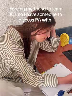 A young woman sits at a desk with a notebook and pen, her head resting on her hand. She's surrounded by a desk setup that includes a laptop, a small yellow fan, and a notepad. The image is cropped, focusing on the woman and the items on the desk with a soft, natural light.