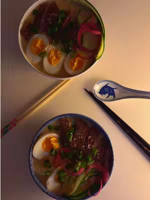 Two bowls of ramen are diagonally arranged on a white surface. The top bowl is slightly tilted, and each bowl is filled with broth, noodles, eggs, sliced vegetables, and meat. Chopsticks and a decorative spoon rest beside the bowls, and the lighting is soft, casting shadows.