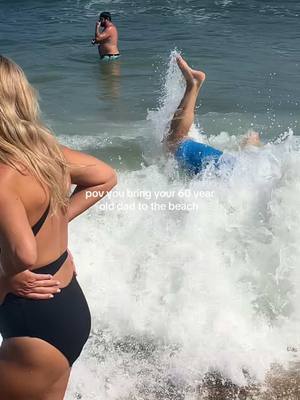 A woman with blonde hair stands on the left side of the frame, looking towards the right. A man in blue shorts is seen upside down in the water with his legs up in the air, creating a splash. In the background, a man is standing in the water.